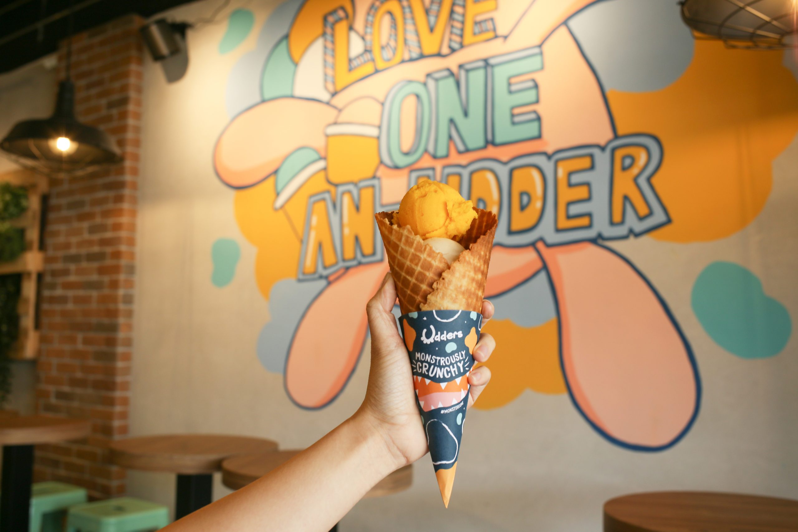 A hand holding an Udders waffle cone with a scoop of mango ice cream in front of a colourful "Love One An-Udder" mural at the Kovan outlet in Singapore