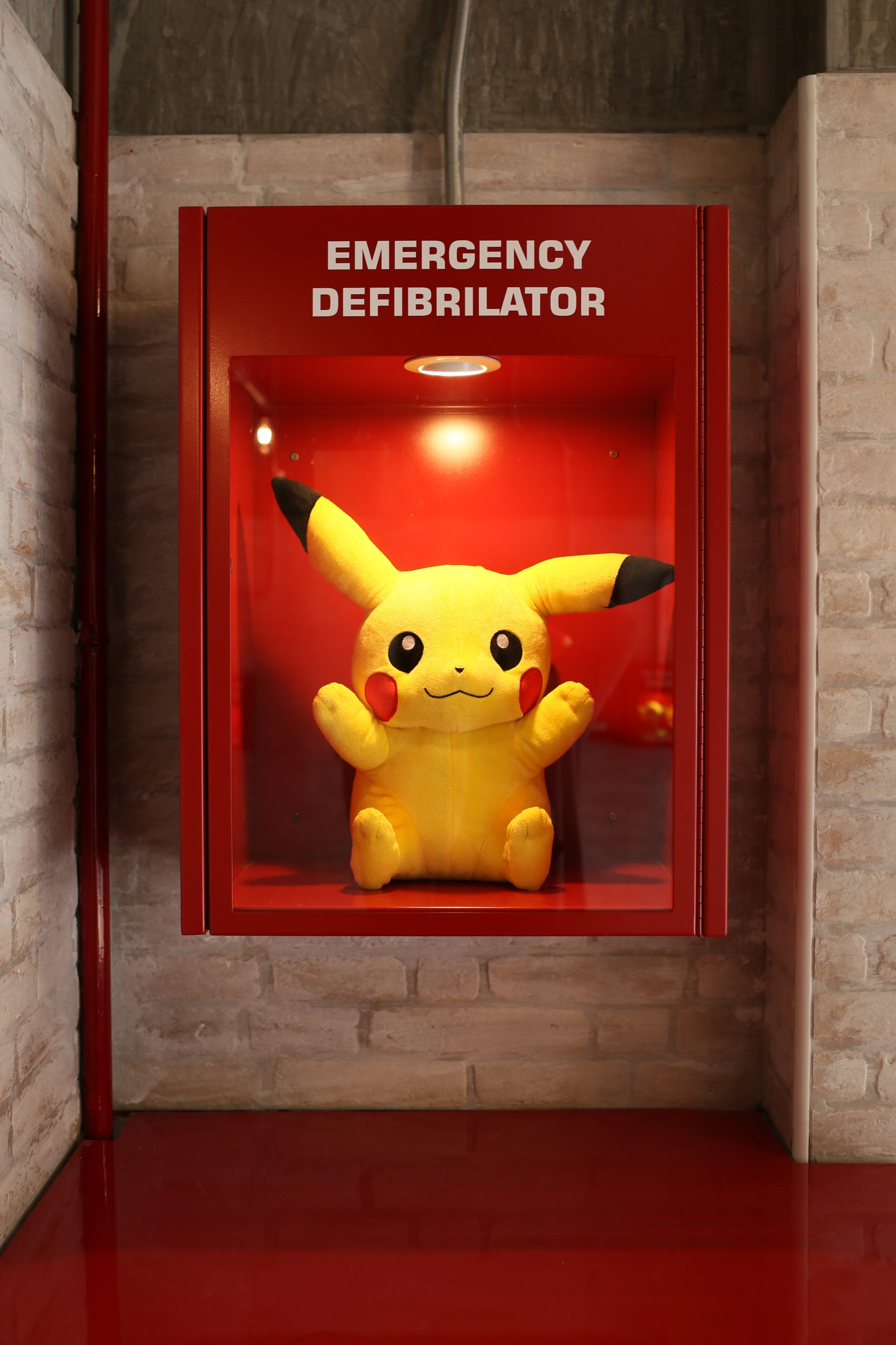 A playful Pikachu plush toy displayed inside a red emergency defibrillator box on the brick wall interior of the Kilat ice cream shop in Singapore
