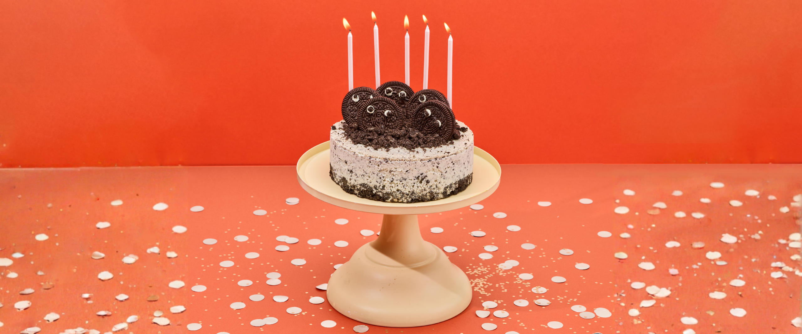 A Udders cookies and cream ice cream cake topped with Oreo cookies and lit birthday candles, surrounded by confetti on a cake stand