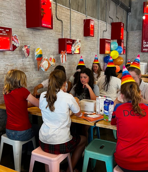 A group of girls in birthday hats celebrating and making ice cream together at an Udders party in Singapore, with colourful balloons and birthday bunting in the background